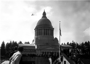 A small airplane flies above a capitol building dome while large crowds look on. 